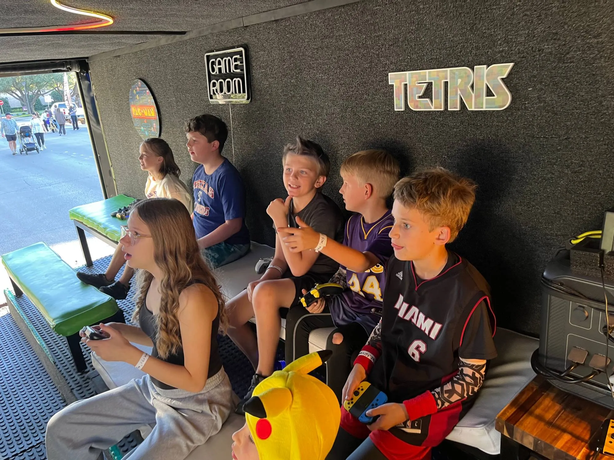 Kids playing video games on the benches inside the Green Games trailer, under the Game Room and Tetris signs